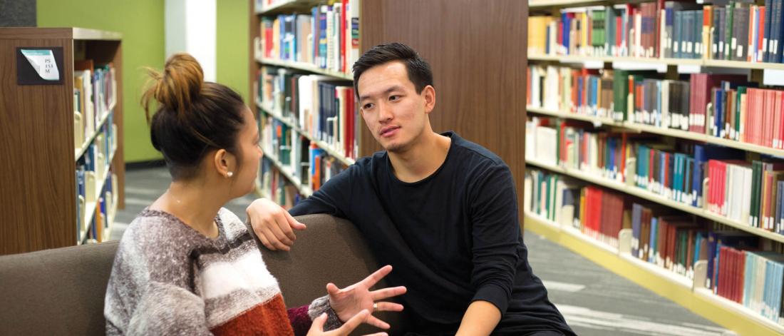 Two students sitting and talking together in a library. 