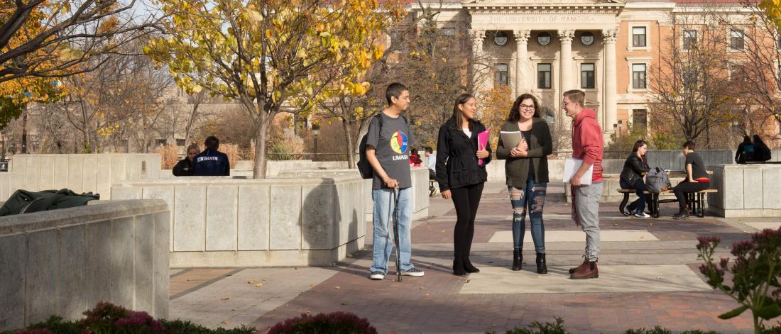 Four students stand together outside of University Centre, several other students are sitting at picnic tables in the background. 