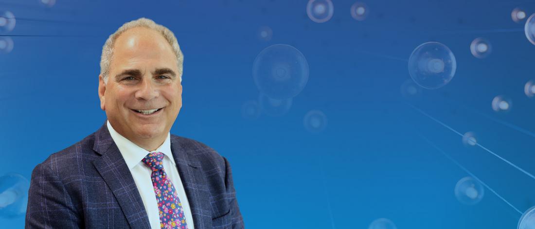Portrait of Lorrie Kirshenbaum smiling in a suit and floral tie, set against a blue background with abstract circular light elements.