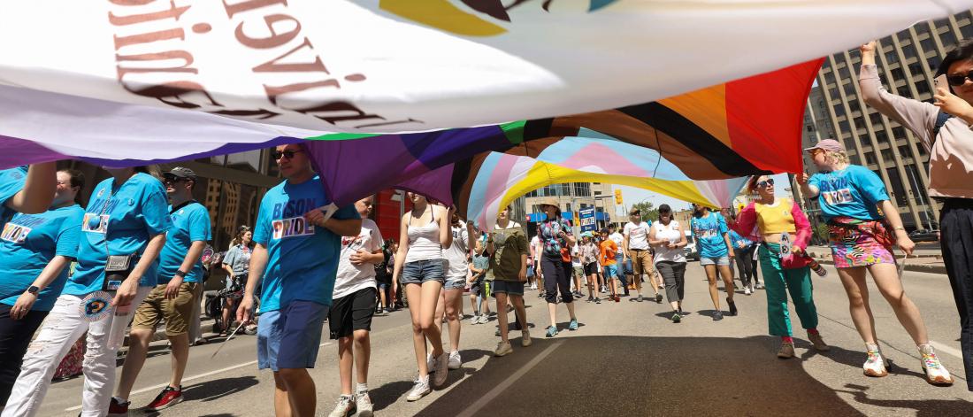 Participants march in a Pride parade carrying a large rainbow and progress pride flag overhead, with several people wearing Bisons Pride shirts.