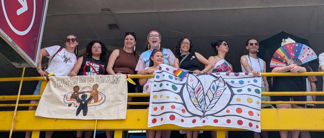 A group of 2ST participants at the Pride Parade standing in a parking garage looking over the bright yellow railings while holding two 2ST banners.