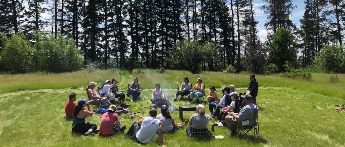 A group of people sitting in a circle in the grass with large trees in the background.
