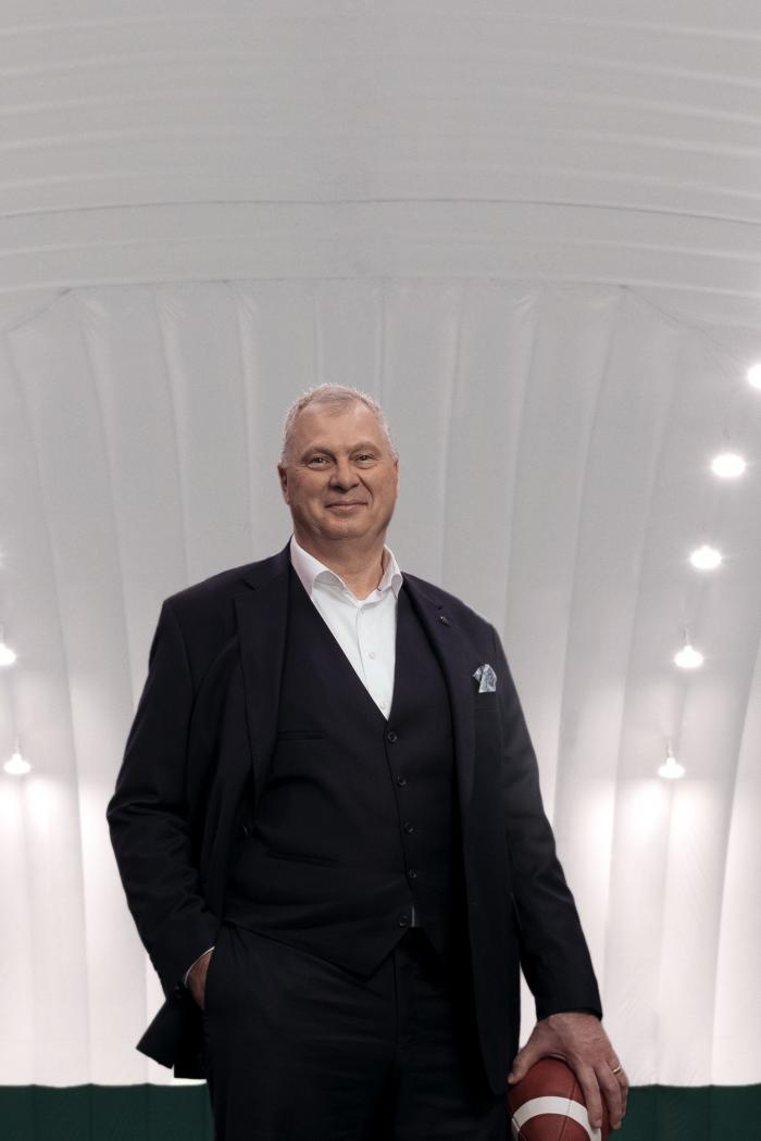 Randy Ambrosie is shown in a dark suit holding a football in his hand and standing on an indoor-football field.