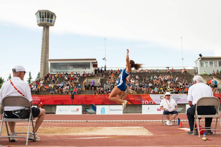 A competing long jump athlete caught mid air over the sand pit while a crowd of spectators watch from the University Stadium stands.