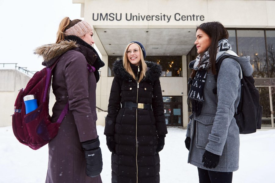 Three young women stand together outside of the UMSU University Centre building.
