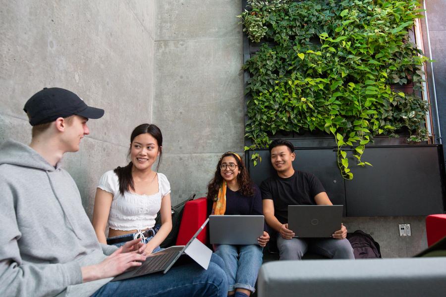 An image of four students lounging by a green wall with laptops conversing with each other and laughing.