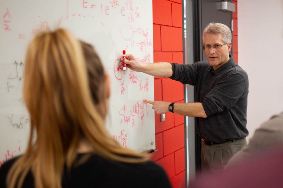 An instructor stands at a dry erase board writing equations in a red marker as a student looks on in the foreground.