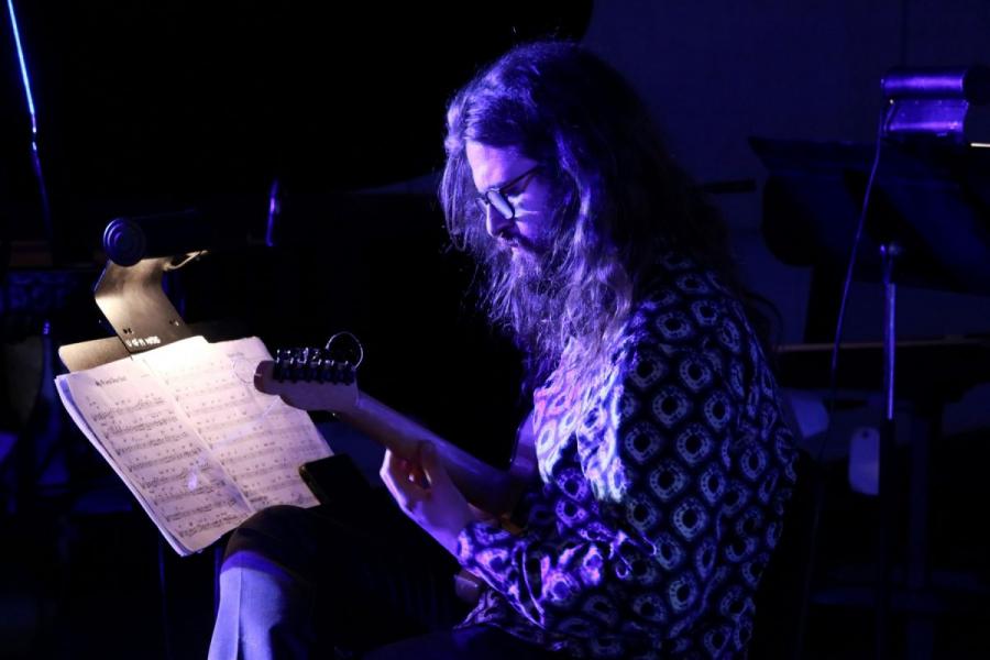 A musician with long hair and a beard plays the guitar in blue lighting in front of sheet music.