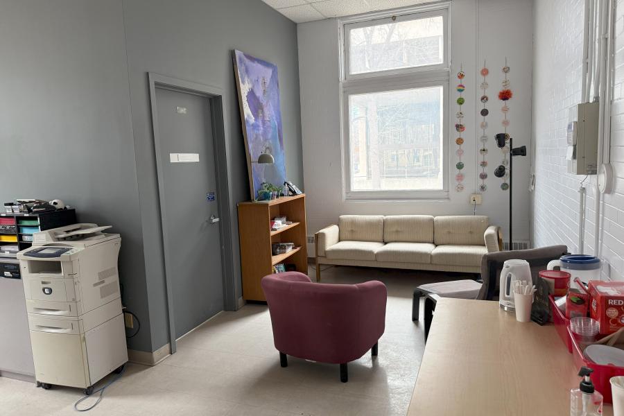 A sitting area with a white couch and red chair. There is one large purple and blue painting above a bookshelf on the left, some art on ribbon next to the window, and a black lamp in the corner. 