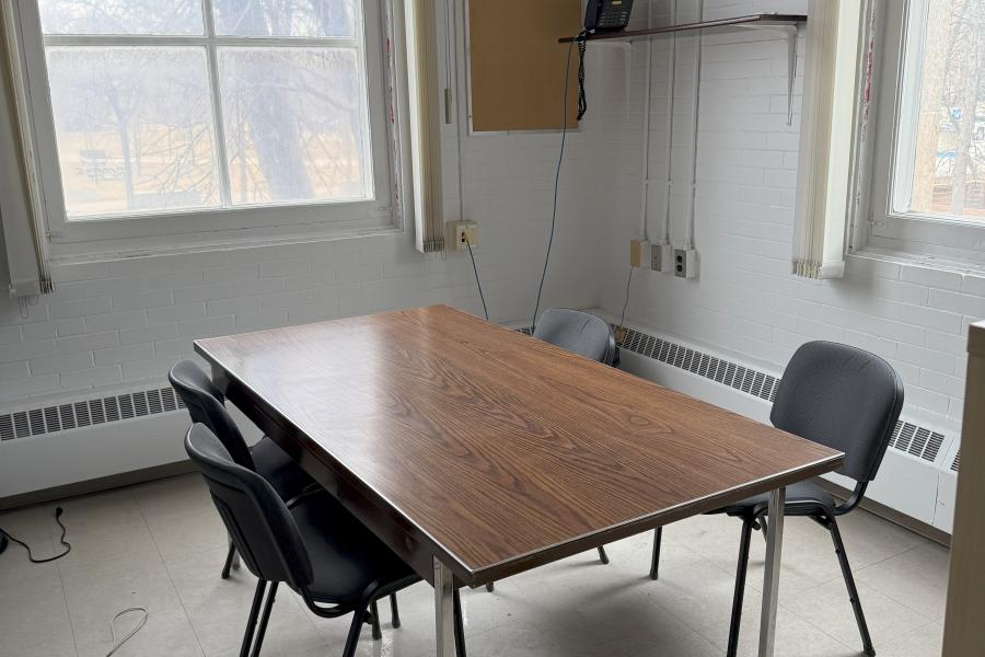 A small room with a brown, rectangular table in the centre and four chairs around it. There is a large window in the back wall and the right wall. 