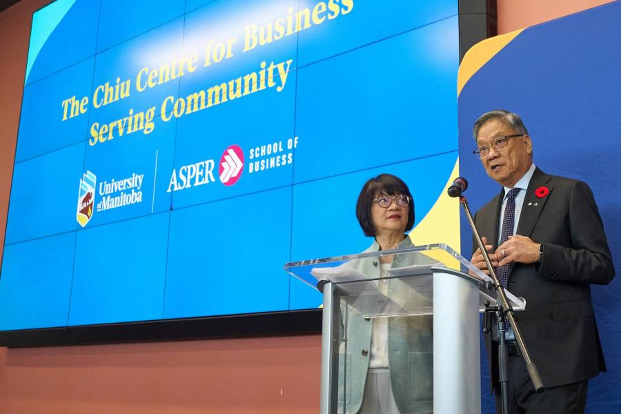 Two speakers stand at a podium in front of a large screen displaying the launch of the Chiu Centre for Business Serving Community at the University of Manitoba’s Asper School of Business.
