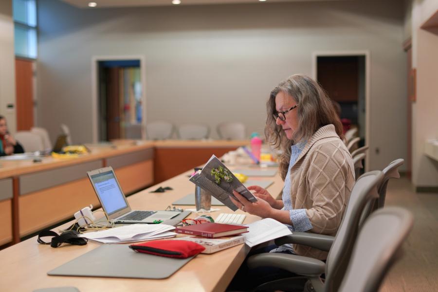 Student looking at a book, sitting in large room with a laptop and writing materials on the table.