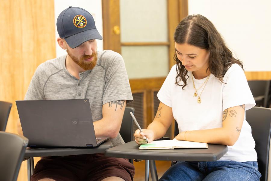 Man with hat sitting at a laptop to the left of a woman writing in a notebook in a classroom setting.