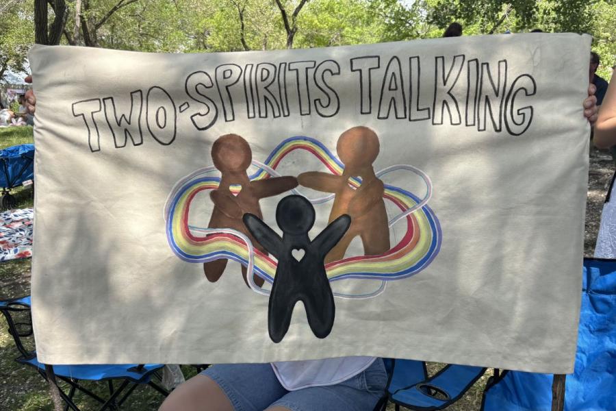 A cream coloured fabric banner consisting of three abstract human figures, in various shades, holding hands in a circle with a heart shape on their chest. The trio is encircled by flowing lines in the trans and rainbow pride flag colours, and above them reads “Two-Spirits Talking”.