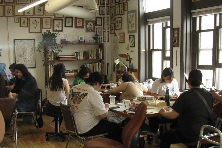A well lit room with groups of 3-4 people sitting around tables. The people are looking at the table in front of them writing letters.
