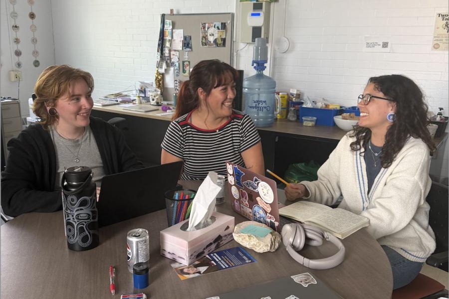 Three people sit at a cluttered table, smiling at one another.