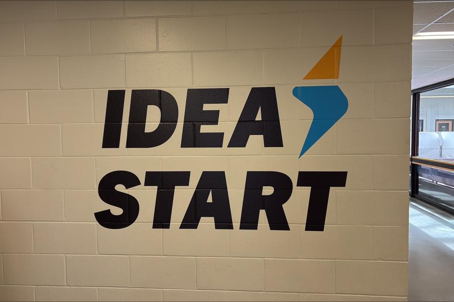 IDEA START logo displayed in large black, blue and orange lettering on a white brick wall inside the University of Manitoba.