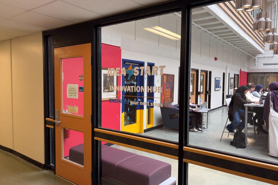 View through the glass entrance of the IDEA START Engineering Innovation Hub, showing students working at tables, colourful doors and lounge seating.