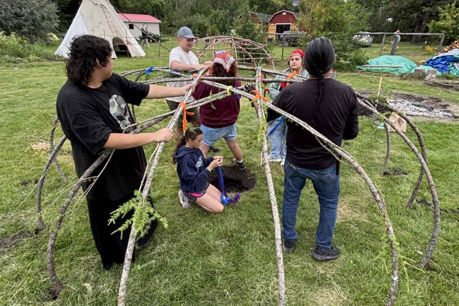 A group of people work together to build a sweat lodge. They are holding and binding together branches to form a dorm structure.
