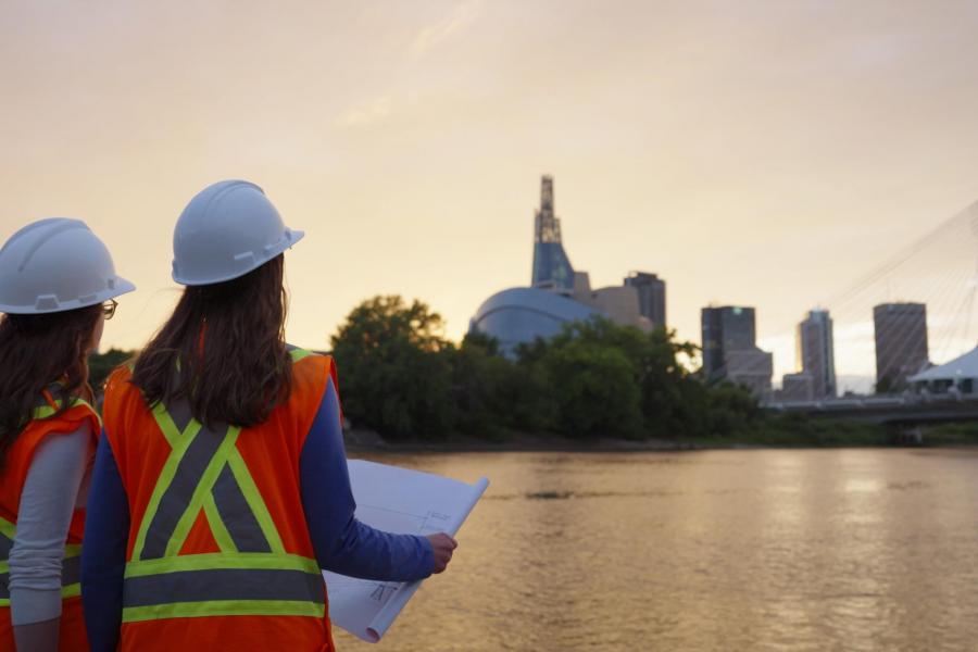 Two people with hardhats hold a technical drawing and look at the Winnipeg skyline.