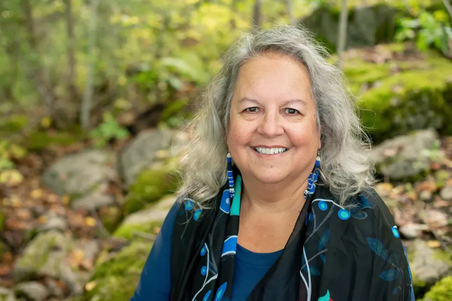 Smiling woman with long grey hair wearing blue beaded earrings and a blue and black patterned scarf, photographed outdoors in a wooded setting with moss-covered rocks.