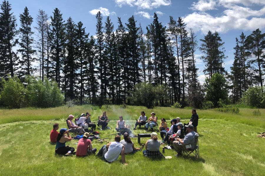 Land and Water group sitting in a circle in a green field with pine trees and blue sky and whisps of clouds in the background