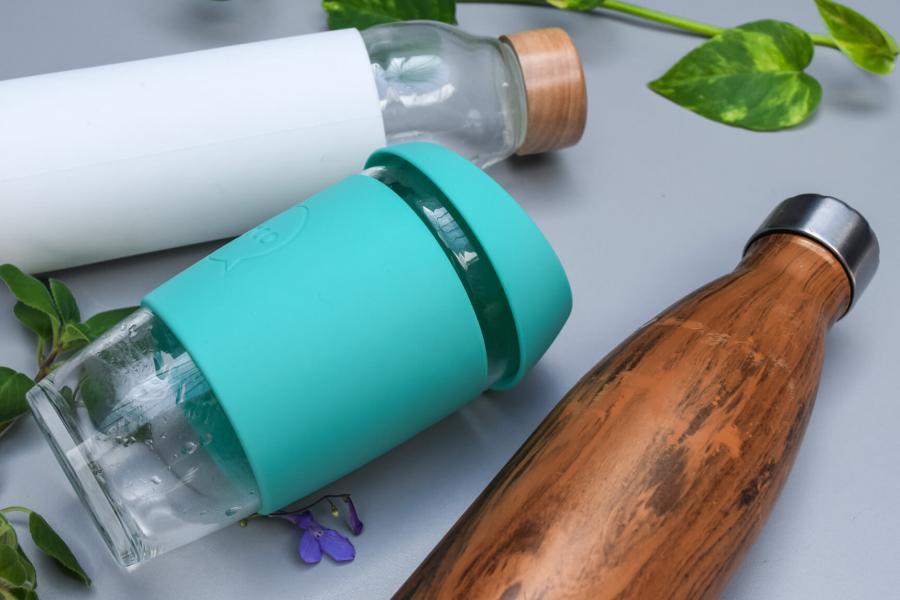 A grey surface with three different re-usable vessels for drinking coffee or water with some plant leaves decoratively laying beside them.